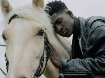 A man wearing a black leather jacket gently embraces and leans against a horse.