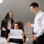 Woman holding HELP sign surrounded by colleagues in a tense office situation.