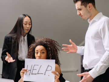 Woman holding HELP sign surrounded by colleagues in a tense office situation.