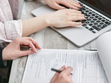Close-up of hands typing on a laptop and reviewing business documents, focused on finance and legal tasks.