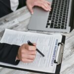 An adult reviews and signs a home insurance policy document on a clipboard next to a laptop.