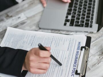 An adult reviews and signs a home insurance policy document on a clipboard next to a laptop.