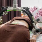 Crop anonymous female patient lying under lasers of fat loss machine on belly during treatment in light modern medical clinic