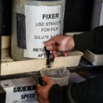 Hands pouring fixer from a jug into a measuring cup for film development.