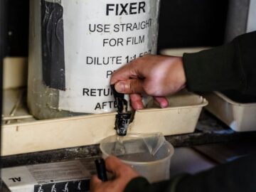 Hands pouring fixer from a jug into a measuring cup for film development.