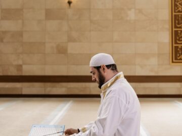 A Muslim man kneels in prayer, reading the Quran inside a mosque, symbolizing devotion.