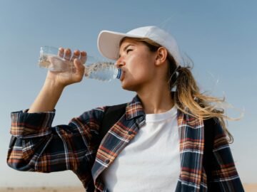 A woman in a baseball cap drinks water from a bottle under a clear blue sky.