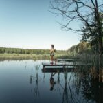 A woman walks on a wooden dock by a calm lake surrounded by trees and clear skies.