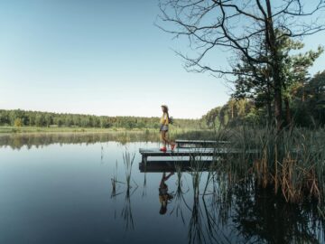 A woman walks on a wooden dock by a calm lake surrounded by trees and clear skies.