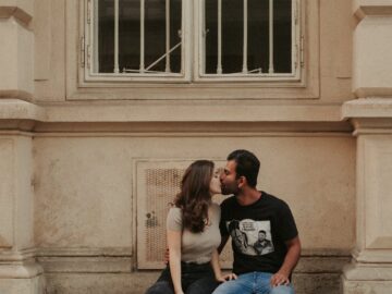 A couple kisses while sitting on a ledge of an urban building facade, expressing love and tenderness.