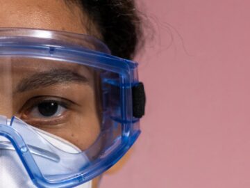 Detailed close-up of a woman wearing safety goggles and a mask against a soft pink background.