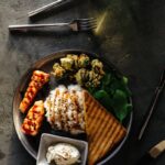 A gourmet dish featuring grilled salmon, rice, greens, and toast on a dark table setting.
