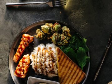 A gourmet dish featuring grilled salmon, rice, greens, and toast on a dark table setting.