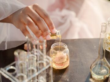 A woman in a pink dress pours liquid into a jar outdoors, surrounded by test tubes.