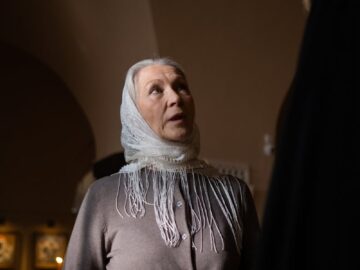 An elderly woman wearing a headscarf converses with a bearded man in a dimly lit room.