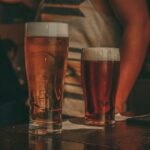 Close-up of two pints of beer on a bar counter in a lively pub setting.