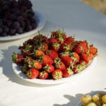 Vibrant strawberries on a white plate surrounded by grapes in natural sunlight.