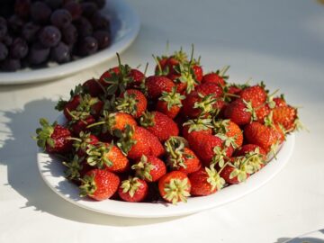 Vibrant strawberries on a white plate surrounded by grapes in natural sunlight.