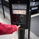Person pressing pedestrian crossing button at street intersection in urban environment.