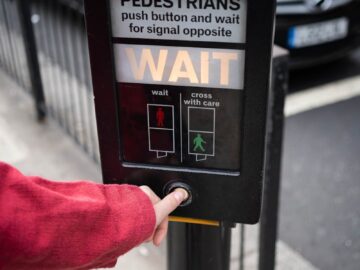 Person pressing pedestrian crossing button at street intersection in urban environment.