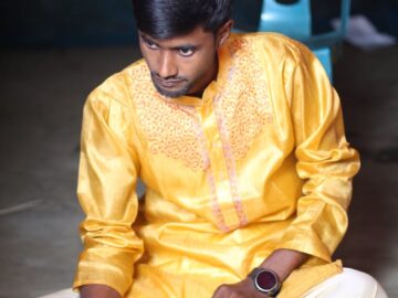 A man in a yellow kurta sits cross-legged indoors, engaging in a traditional ritual.