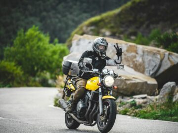 A motorcyclist rides through a scenic mountain road, making a peace sign.