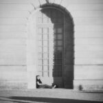 A person relaxes under a large archway against a sunlit wall, casting long shadows.
