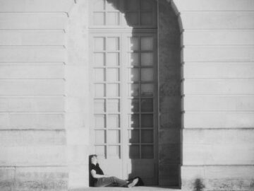 A person relaxes under a large archway against a sunlit wall, casting long shadows.