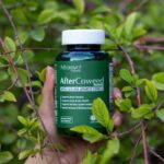 Hand holding a supplement bottle amidst vibrant green foliage outdoors.