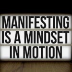 Inspirational quote 'Manifesting is a mindset in motion' on lightbox over dark background.