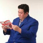Adult man in blue suit reads a magazine against neutral background.