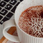 Cozy cup of cocoa with cocoa powder on top beside a keyboard on a work desk.