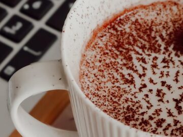 Cozy cup of cocoa with cocoa powder on top beside a keyboard on a work desk.