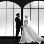 A black and white photo of a couple in wedding attire standing indoors, holding hands.