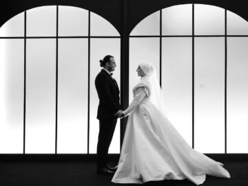 A black and white photo of a couple in wedding attire standing indoors, holding hands.