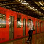 Man holding phone on metro platform beside a brightly colored train.