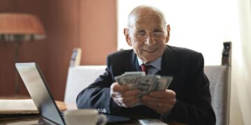 Elderly man smiling while counting cash at a home office setting.