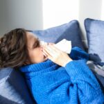 A woman in a blue sweater uses a tissue, lying indoors, suggesting illness or flu symptoms.
