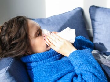A woman in a blue sweater uses a tissue, lying indoors, suggesting illness or flu symptoms.