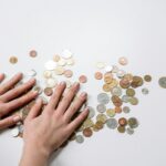 Hands arranging a variety of international coins on a neutral light background.