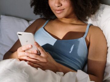 African American woman enjoying a cozy morning in bed, using a smartphone.