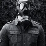 Black and white faceless male in rubber gas mask and casual shirt standing against blurred foliage