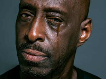 A close-up portrait of a man shedding a tear against a gray background, depicting sadness and contemplation.