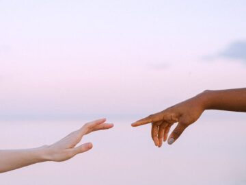 Two diverse hands reaching towards each other over a serene pastel sky and water background.