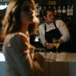 Stylish woman at a bar counter with a bartender in a dimly lit, elegant setting.