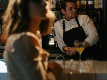 Stylish woman at a bar counter with a bartender in a dimly lit, elegant setting.