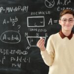 Young boy holding chalk in front of blackboard with math formulas, wearing glasses and smiling.