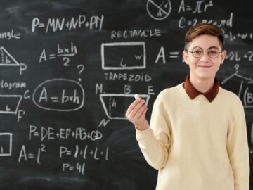 Young boy holding chalk in front of blackboard with math formulas, wearing glasses and smiling.