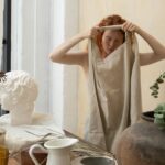 Young female artisan with closed eyes putting on apron near table with jug and glassware in workshop