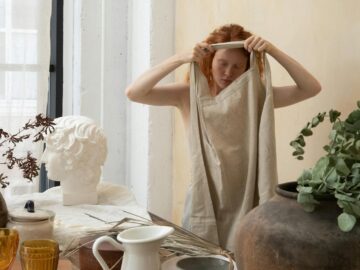 Young female artisan with closed eyes putting on apron near table with jug and glassware in workshop